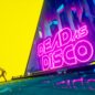 La scritta neon di Dead as Disco che copre la metrà destra dello schermo, mentre sulla sinistra la silhoutte di Disco balla su sfondo giallo